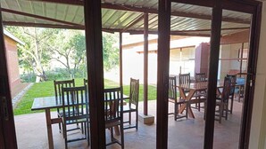 A covered patio area with wooden tables and chairs, a concrete floor, and a view of a grassy area and buildings in the background. - Chiyedza Place (Juliasdale)
