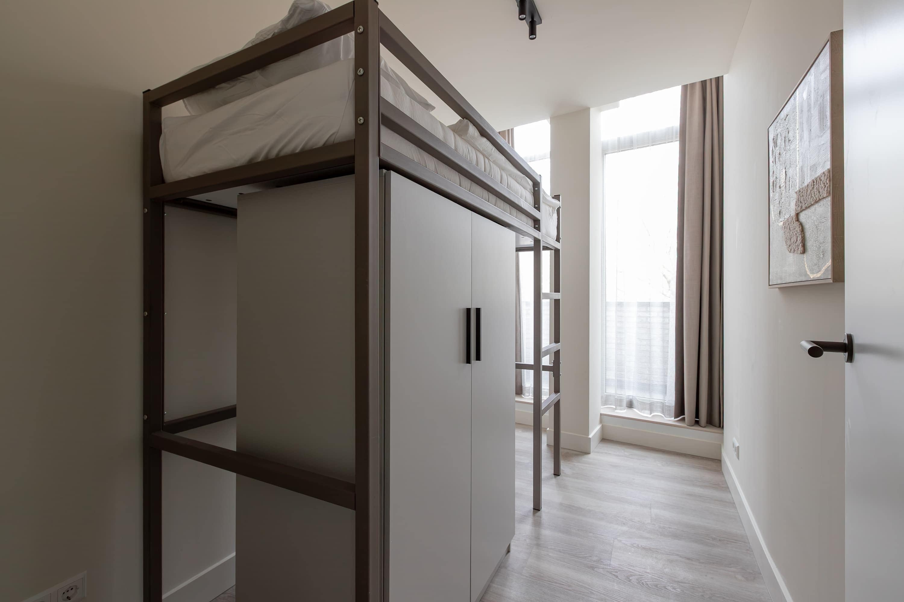 A bunk bed with a ladder, a white wardrobe, a door, and a window with curtains.