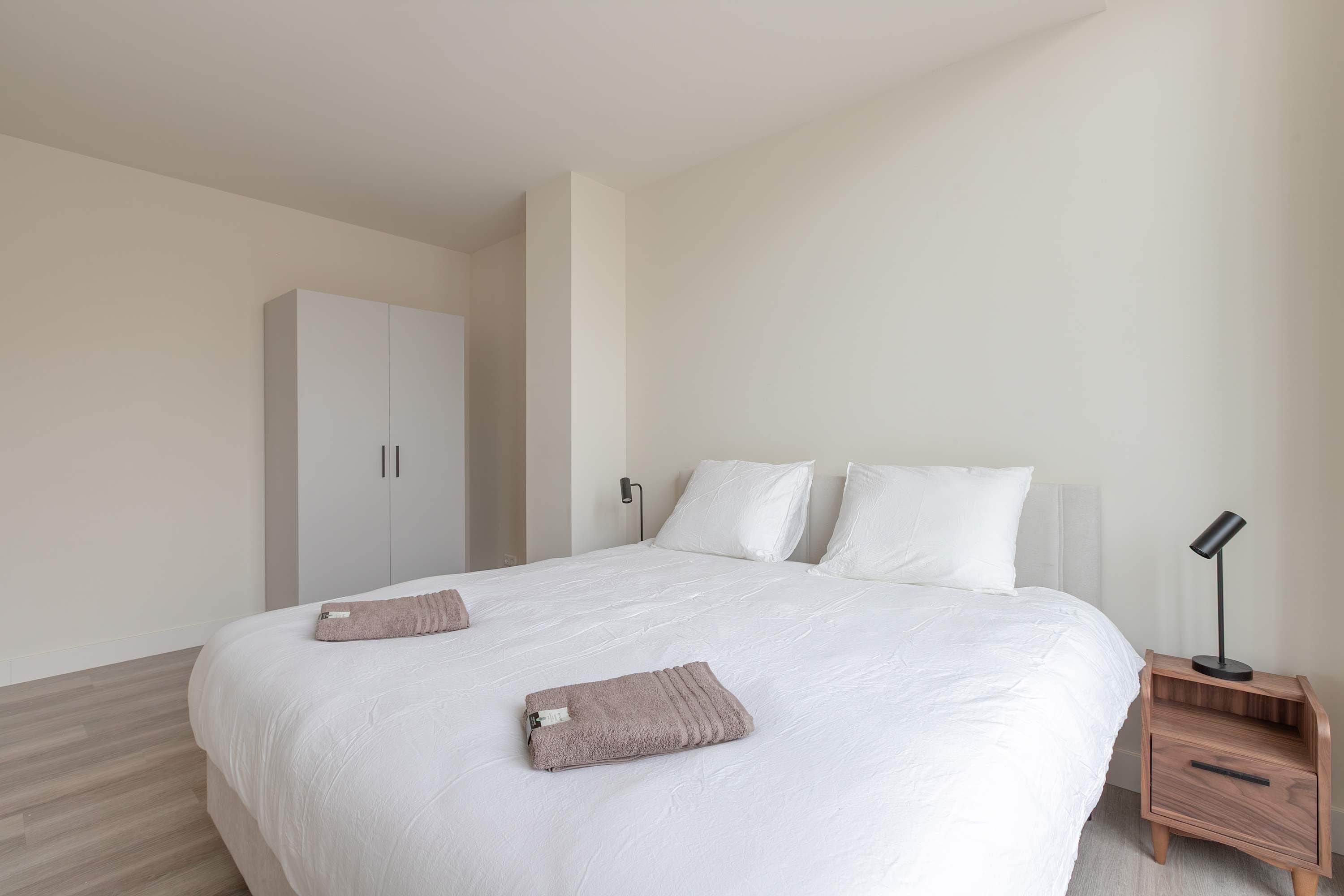 A bedroom with a large bed, a wardrobe, a nightstand with a lamp, and a wooden floor.