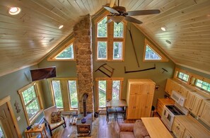 A loft-style room with a stone chimney, wooden beams, a ceiling fan, and large windows.