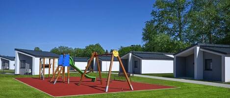 Un parque infantil con columpios y toboganes, rodeado de edificios modernos con techos planos.