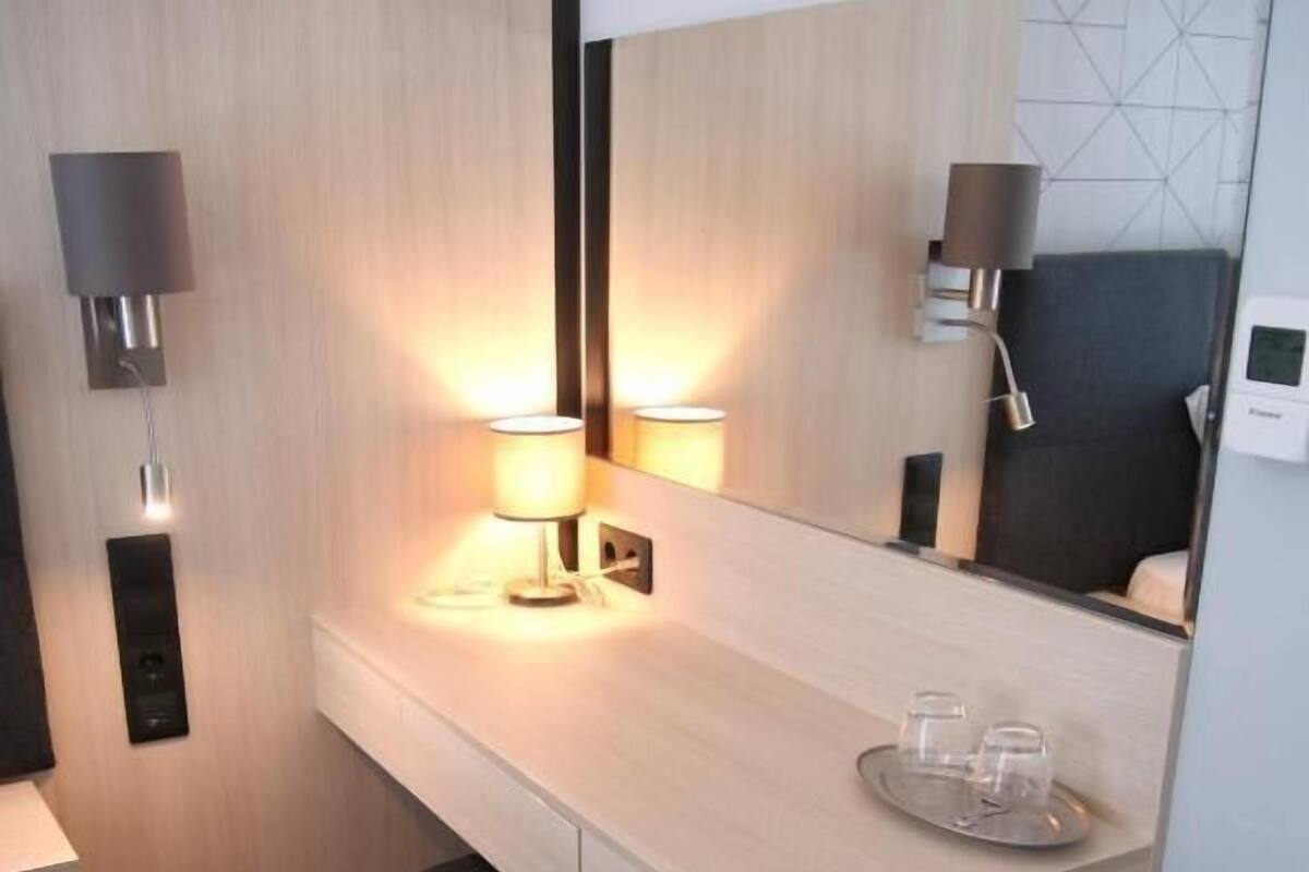 A hotel room with a wooden desk, two wall-mounted lamps, a mirror, and a small stool.