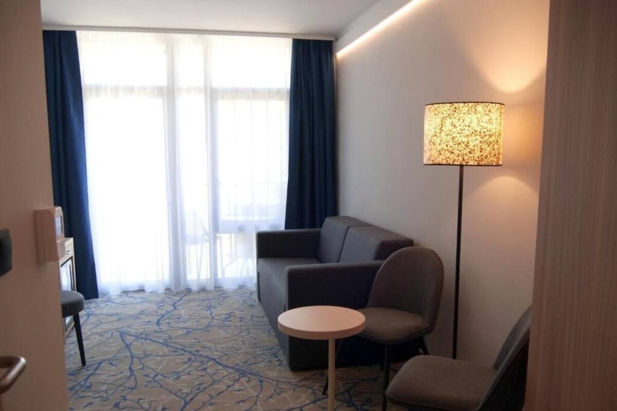 A hotel room with a sofa, two armchairs, a small round table, a lamp, and a large window with curtains.