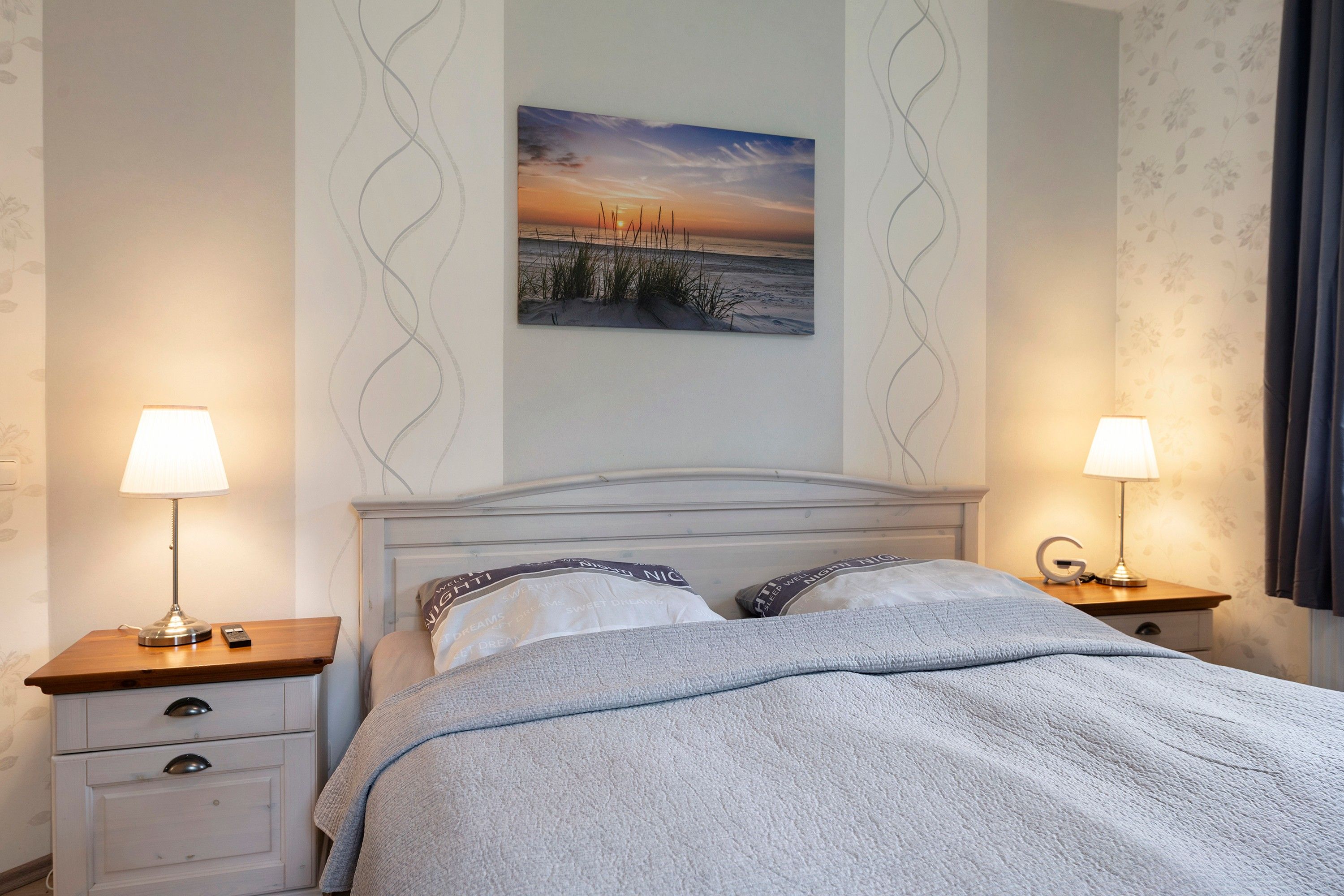 Ein Schlafzimmer mit einem Bett, zwei Nachttischen, einer Nachttischlampe, einem Wandbild und einer Wandtapete mit Muster.