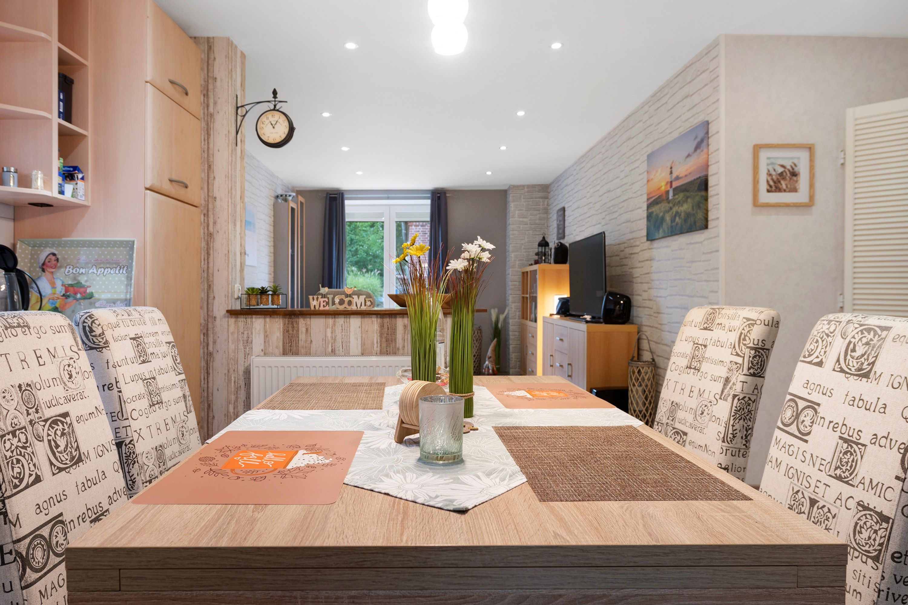 Ein Essbereich mit einem Holztisch, gemusterten Stühlen, einer Blumenvase und einer Uhr an der Wand.