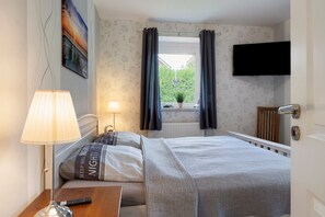 A bedroom with a bed, a bedside table with a lamp, a TV mounted on the wall, and a window with curtains. - Apartment '3 Ebenerdig' (Cuxhaven)
