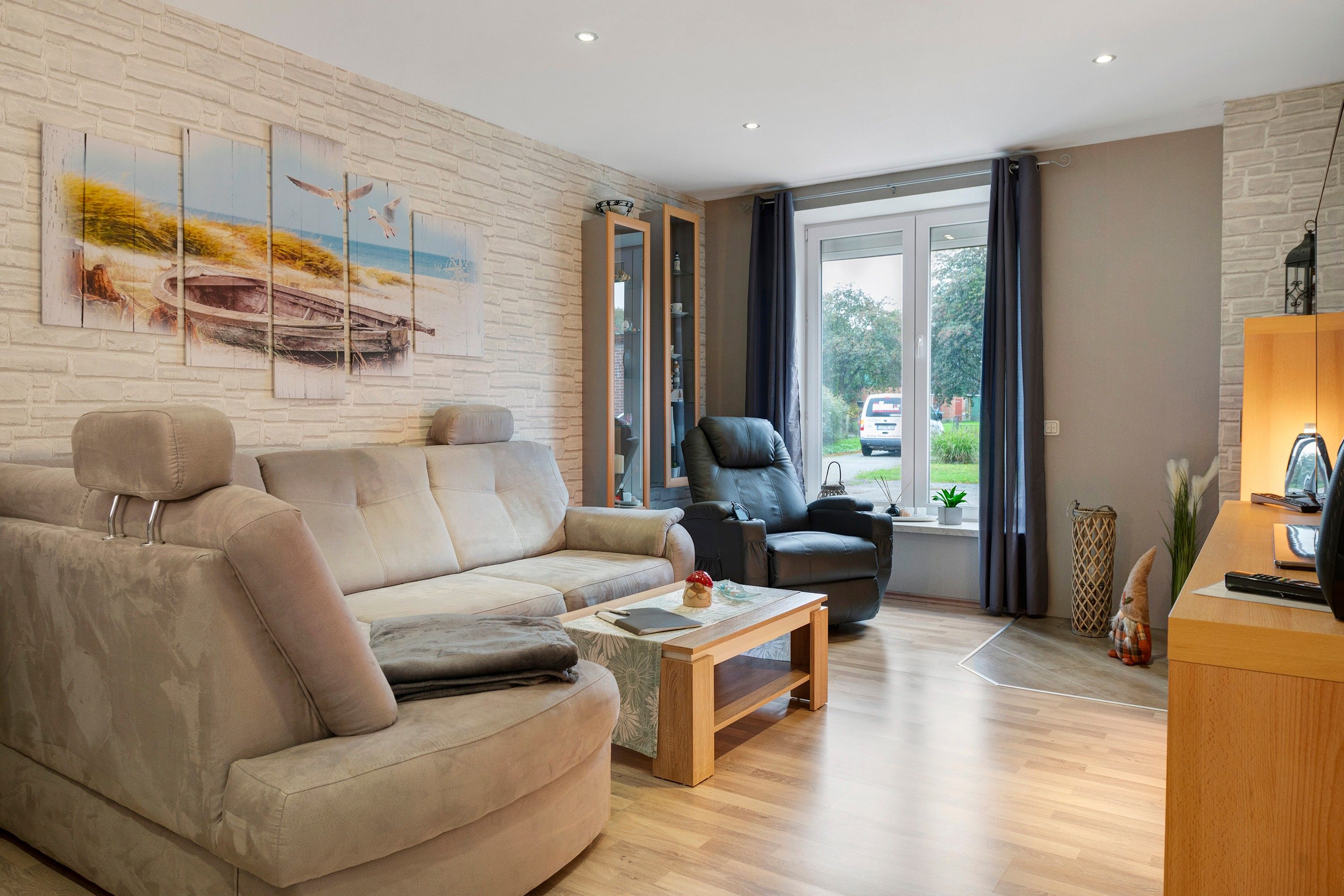 Ein modernes Wohnzimmer mit einem beige-farbenen Sofa, einem schwarzen Ledersessel, einem Holztisch und einem großen Fenster mit Vorhängen.