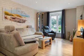 A modern living room with a beige sofa, a black leather chair, a wooden coffee table, and a large window with curtains. - Apartment '3 Ebenerdig' (Cuxhaven)