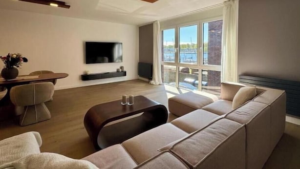 Ein modernes Wohnzimmer mit Sofa, Couchtisch, Wandfernseher und großen Fenstern mit Blick auf das Wasser.