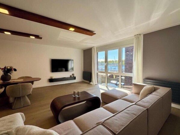 A modern living room with a sofa, a coffee table, a TV mounted on the wall, and large windows with a view of the water. - Bayfront Escape - Stylish time out by the water in Travemünde (Lübeck)