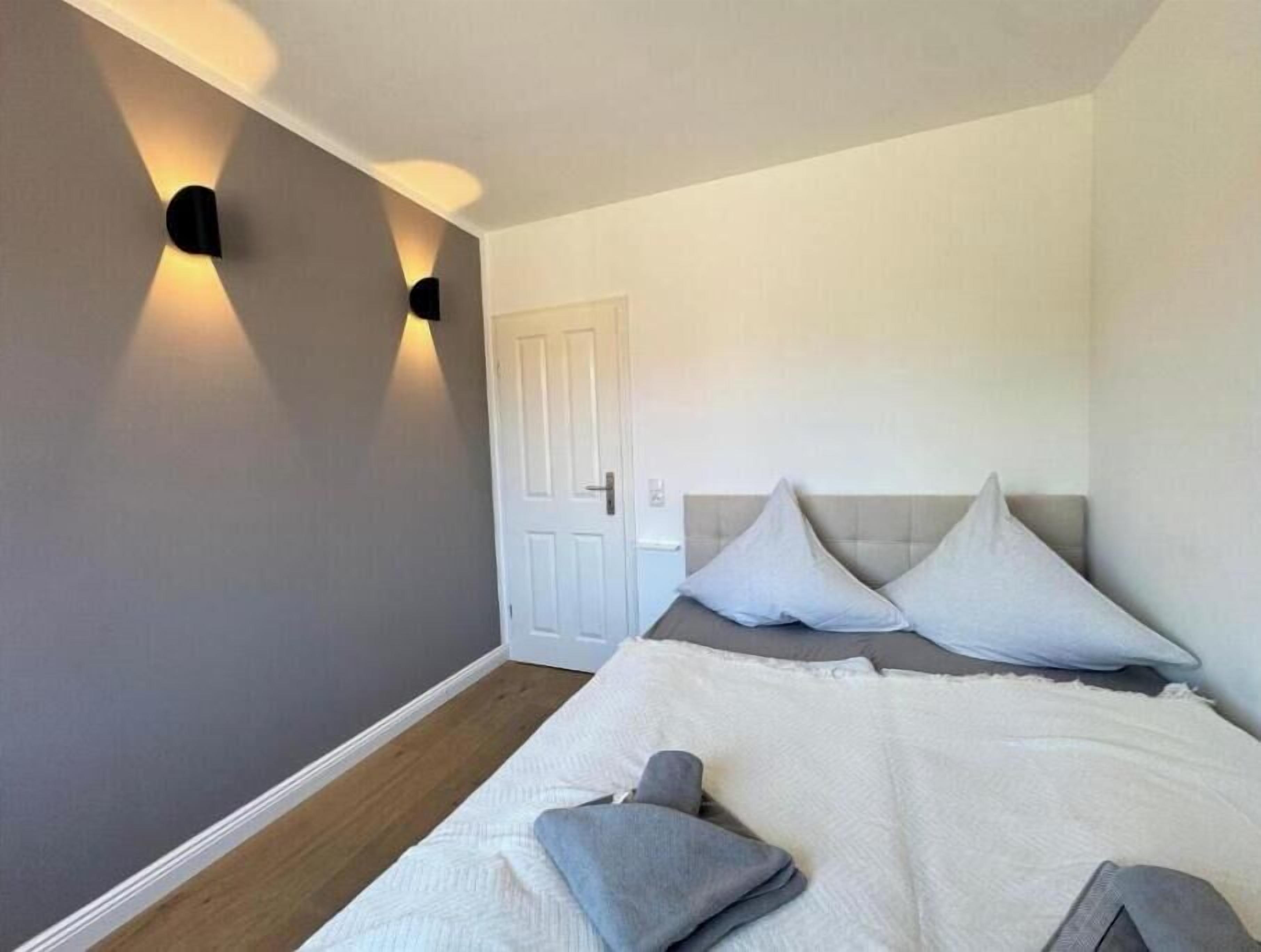 A bedroom with a bed, pillows, a nightstand, a wall-mounted light, and a door.