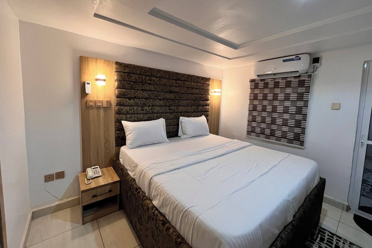 Una habitación de hotel moderna con una cama grande, cabecera de madera, lámparas de noche, teléfono y equipo de aire acondicionado.