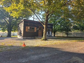 Ein kleines Gebäude mit Holztür und -fenstern, umgeben von Bäumen und einem Kiesplatz.