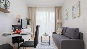 A compact living space with a dining table, chairs, a sofa, a TV, and a small side table with a lamp. - Comfortable Apartment | Parking (Warszawa)