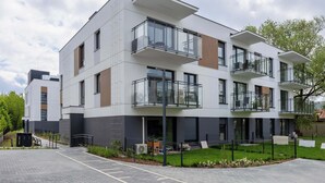 A modern multi-story residential building with balconies, a paved driveway, and a small garden area. - Comfortable Apartment | Parking (Warszawa)