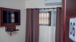 A room with a TV mounted on the wall, a bed with a red and beige bedspread, and a window with curtains. - résidence hoteliere lauria (Lomé)