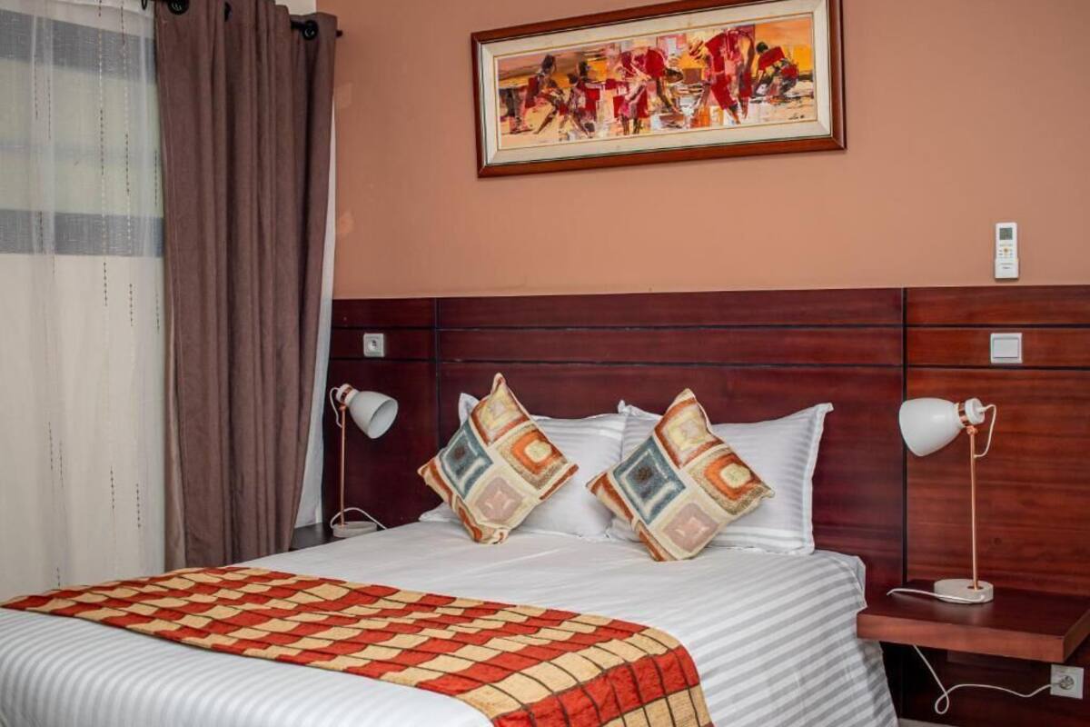 Habitación de hotel con cama, dos lámparas de noche, una mesita de noche, un cuadro en la pared y una ventana con cortinas.
