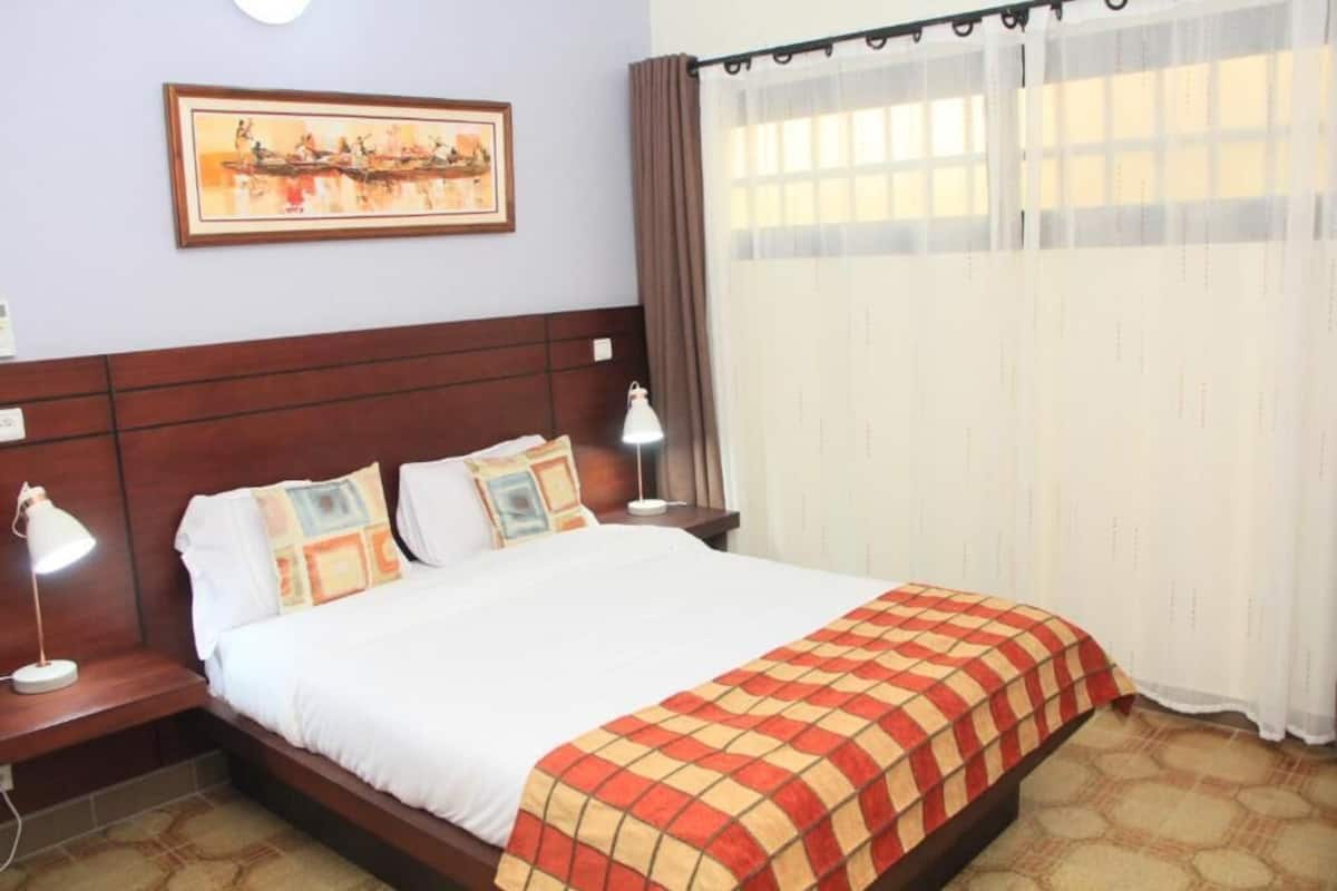 Una habitación de hotel con cama, dos lámparas de noche, cabecera, un cuadro en la pared y ventana con cortinas.