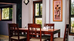 A dining area with a wooden table and chairs, framed artwork on the wall, and windows with views of greenery. - résidence hoteliere lauria (Lomé)