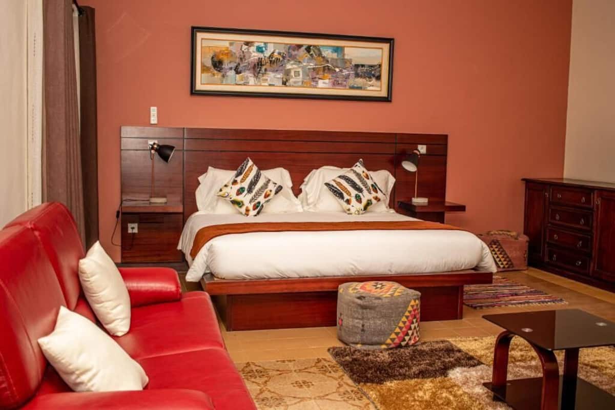 Un dormitorio con cama, sofá rojo, pequeño reposapiés, mesita de noche, lámpara, cómoda y un cuadro en la pared.