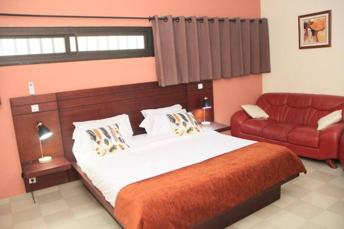 Un dormitorio con cama, un sofá rojo, una mesita de noche con lámpara y una ventana con cortinas.