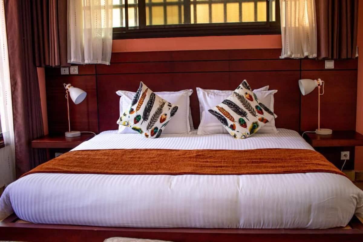 Una cama con ropa de cama blanca, dos almohadas con estampado de plumas, un plaid marrón y una ventana con cortinas.