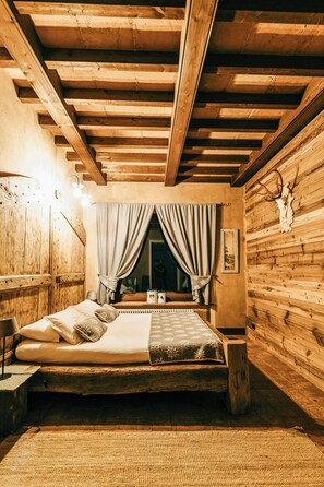 Ein rustikales Zimmer aus Holz mit einem Bett, Holzwänden, einem Fenster mit Vorhängen und einem aufgehängten Tierkopf an der Wand.