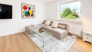 A modern living room with a sectional sofa, a glass coffee table, a flat-screen TV, a colorful abstract painting, and a large window with a view of greenery. - Sea Studio - Modern time-out right on the beach in Scharbeutz (Scharbeutz)