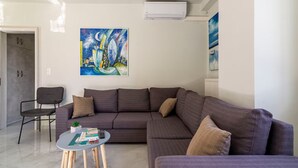 Ein modernes Wohnzimmer mit einem Ecksofa, einem kleinen runden Tisch und einem Sessel. Ein Gemälde hängt an der Wand und ein Klimagerät befindet sich über dem Sofa.
