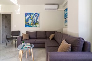Ein modernes Wohnzimmer mit einem Ecksofa, einem kleinen runden Tisch und einem Sessel. Ein Gemälde hängt an der Wand und ein Klimagerät befindet sich über dem Sofa.