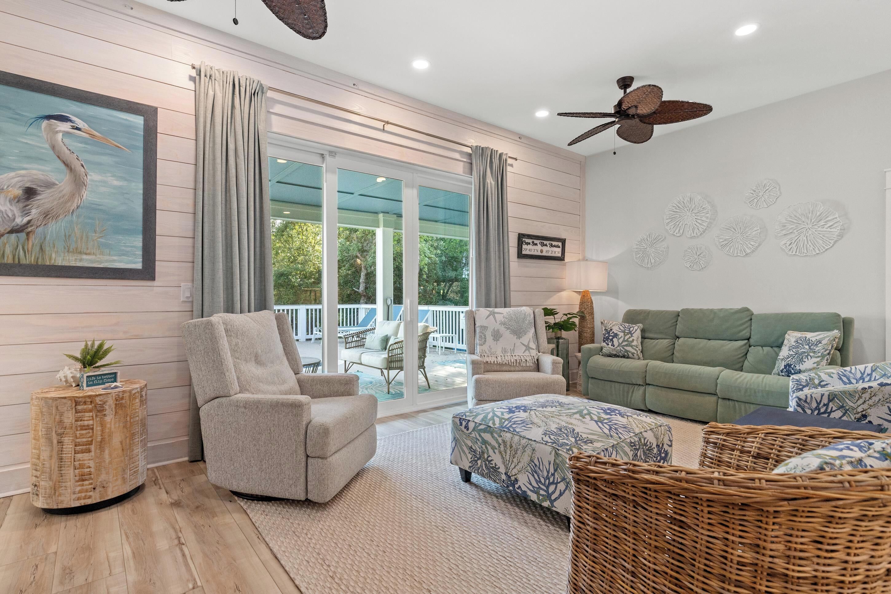 A living room with a green sofa, two armchairs, a wicker ottoman, a ceiling fan, and a large window with curtains.