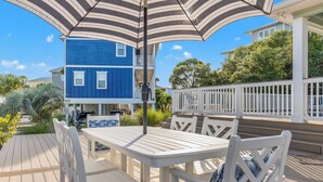 A deck with a table and chairs, a striped umbrella, and a blue house in the background. - Beach House - Coastal Cabana (Port St. Joe)