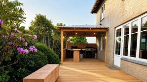 Une terrasse en bois avec un espace barbecue, un banc et une maison dotée de grandes fenêtres.