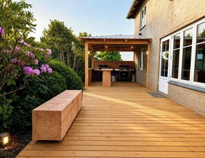 Una terraza de madera con una zona de barbacoa, un banco y una casa con ventanales grandes.