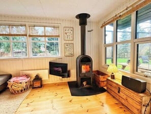 Ein gemütlicher Raum mit Kaminofen, Fernseher und einem großen Fenster mit Blick ins Grüne.