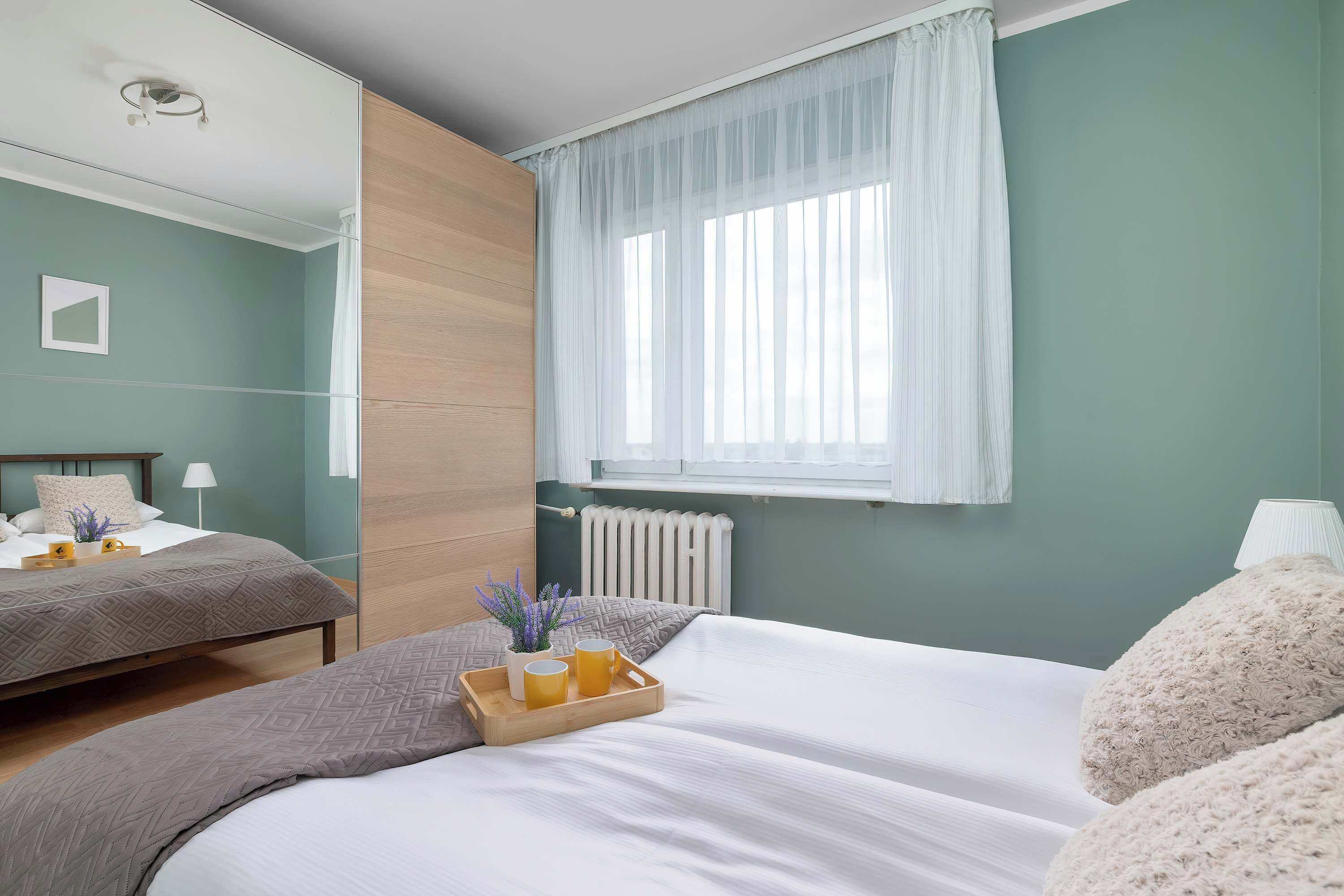 A bedroom with two beds, a window with sheer curtains, a bedside table with a lamp, and a radiator.