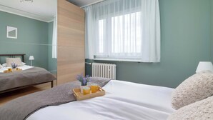 Una camera da letto con due letti, una finestra con tende leggere, un comodino con una lampada e un termosifone.