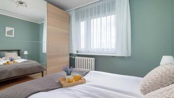 A bedroom with two beds, a window with sheer curtains, a bedside table with a lamp, and a radiator.