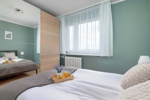 Ein Schlafzimmer mit zwei Betten, einem Fenster mit durchscheinenden Vorhängen, einem Nachttisch mit Lampe und einem Heizkörper.