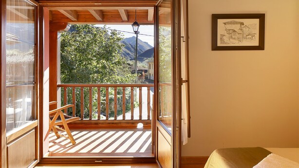 Une pièce avec un plancher en bois, un lit recouvert d’une couverture blanche, un banc en bois et un balcon offrant une vue sur les arbres et les montagnes.