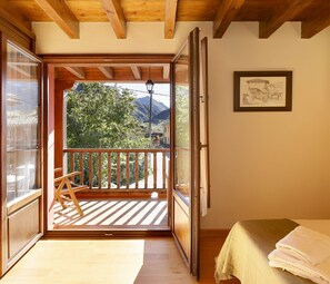Ein Zimmer mit Holzboden, einem Bett mit weißer Bettdecke, einer Holzbank und einem Balkon mit Blick auf Bäume und Berge.