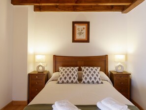 A neatly made bed with two pillows, two bedside tables with lamps, a framed botanical print on the wall, and wooden beams above the bed.