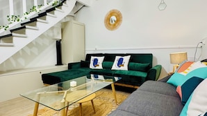 Een moderne woonkamer met een glazen salontafel, een groene bank met gestreepte kussens, een trap met planten en een ronde spiegel aan de muur.