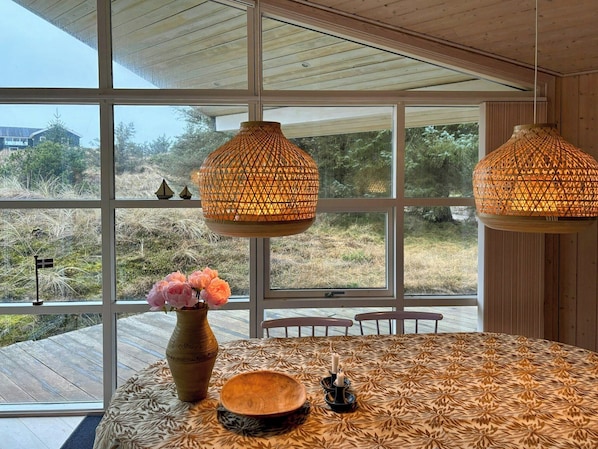 A dining area with a large window, a table with a patterned tablecloth, a vase with flowers, and two hanging lamps. - House with wide views in dunes near Blokhus (Blokhus)