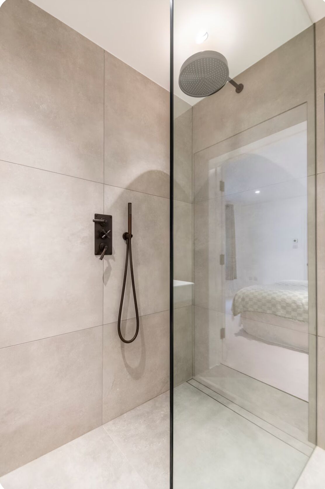 Ein modernes Badezimmer mit Duschkabine aus Glas, einer Handbrause und einem sichtbaren Bett im angrenzenden Zimmer.