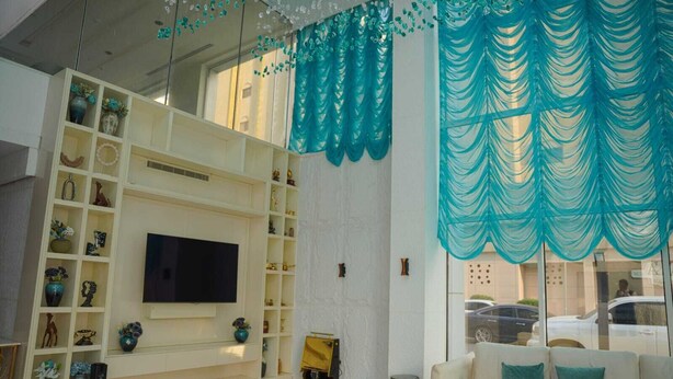 A living room with a flat-screen TV, white shelving with decorative items, a sofa with teal cushions, and teal drapery.