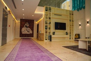 A hotel lobby with a purple carpet, a large wall mural, a TV, and shelves with decorative items. - CASA km 1 serviced apartments - كاسا كي ام ١ للشقق مخدومة (Riyadh)