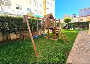 Eine hölzerne Spielkonstruktion auf einer Rasenfläche mit einem gepflasterten Weg und einem Gebäude im Hintergrund.