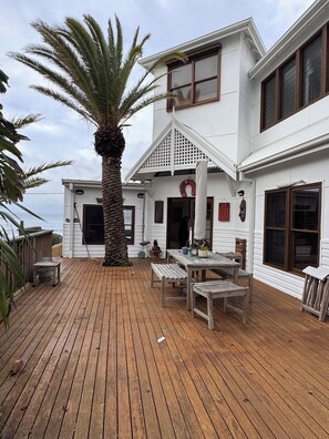 A wooden deck with a palm tree, a covered seating area, and a white building with multiple windows.