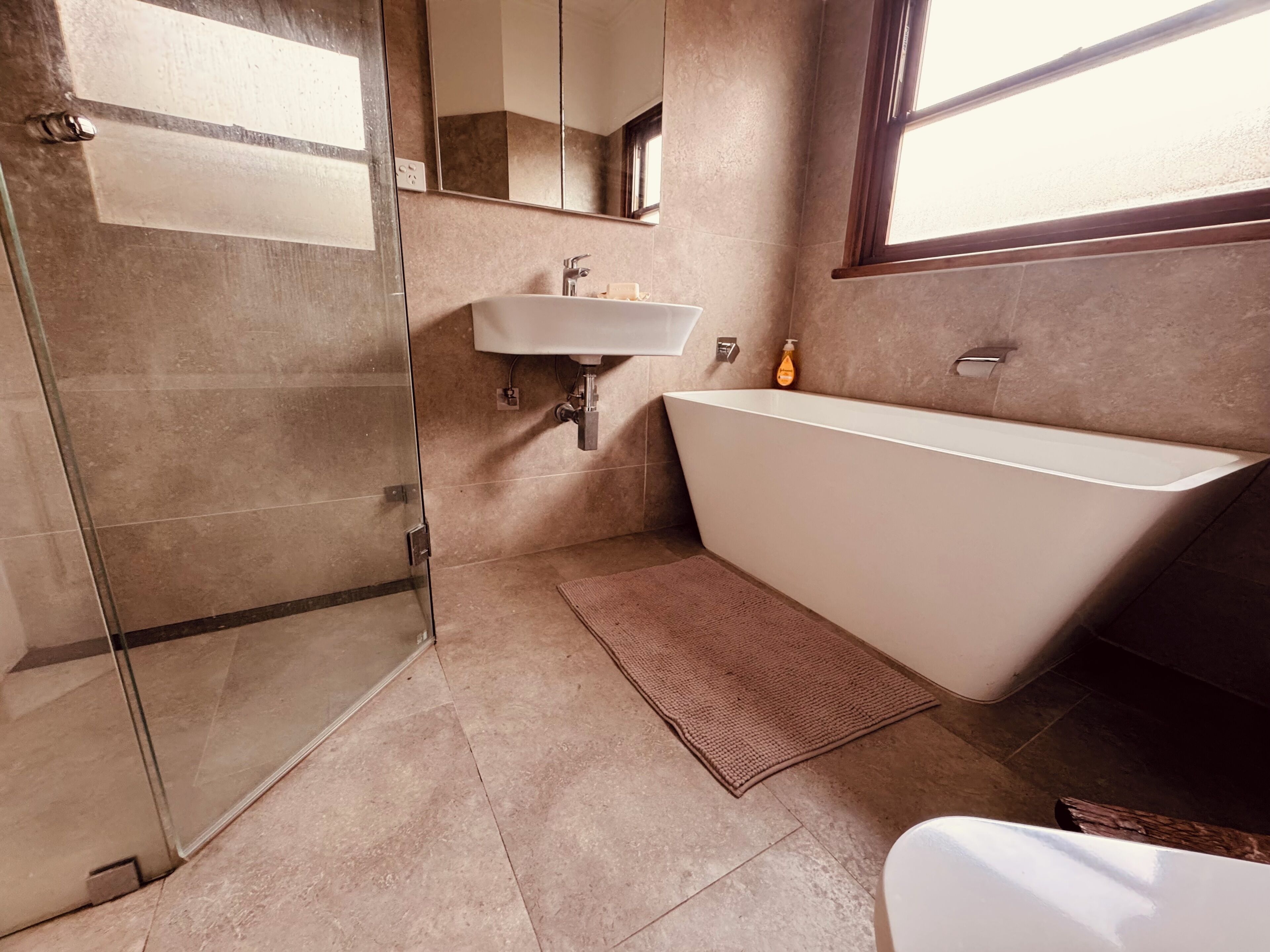 A bathroom with a glass shower enclosure, a freestanding bathtub, a wall-mounted sink, and a window providing natural light.